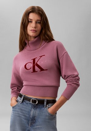 Kvinde med langt brunt hår iført en cropped lyserød højhalset sweater med "CK Calvin Klein" logo og blå jeans med sort bælte.