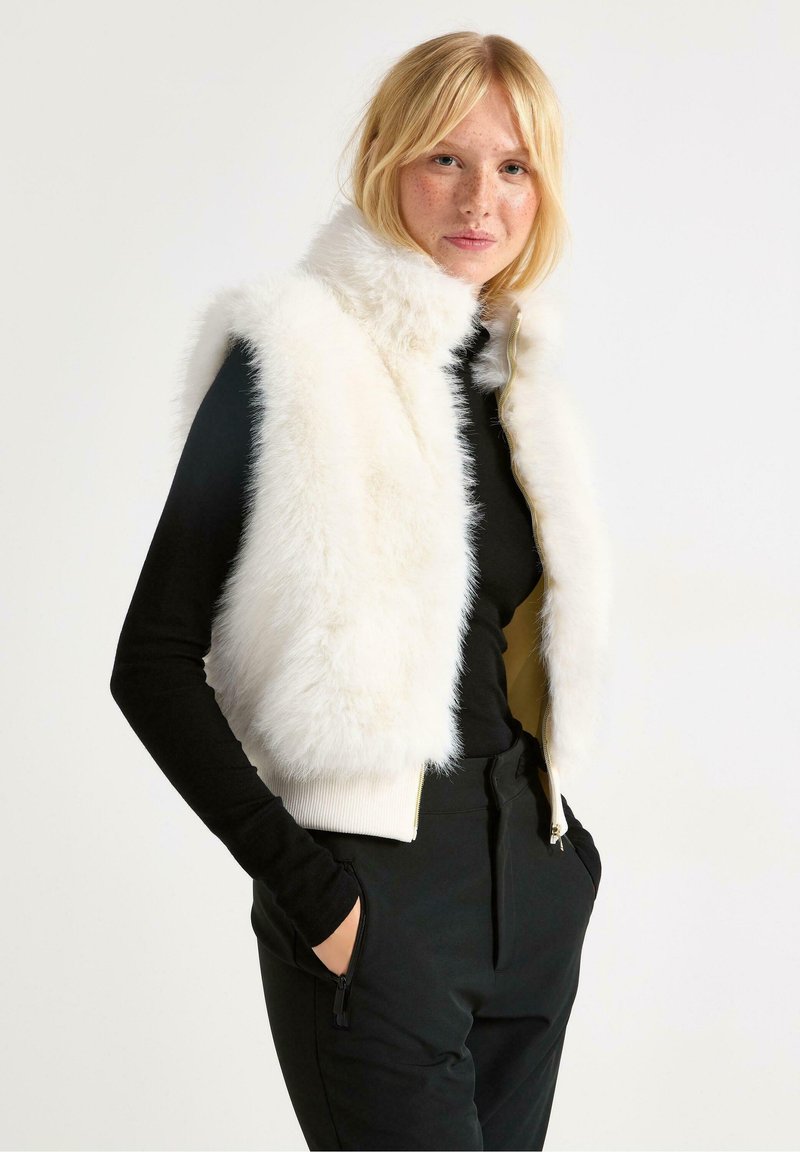 Vesti teko faux vaaleassa värissä, jossa on korkeakauluksinen muotoilu ja kultainen vetoketju, yhdistetty mustaan pitkähihaiseen paitaan ja mustiin housuihin, joissa on vetoketjulliset taskut.