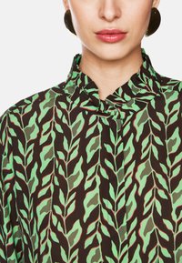 Grüne und schwarze Bluse mit einem Blattmuster, hoher Kragen und gerafftem Ausschnitt. Der Stoff wirkt leicht und fließend.