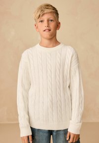 Pullover bianco a maglia cable-knit con colletto rotondo e polsini a costine, indossato sopra jeans in denim blu. La texture è morbida con motivi in rilievo.