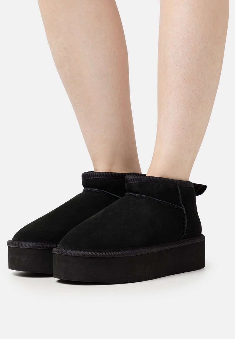 Bottines à plateforme en daim noir portées sur jambes nues, avec des bouts arrondis et une languette à l'arrière, présentées sur un fond blanc.