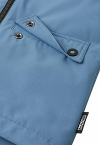Primo piano di un tessuto impermeabile blu con bottoni a scatto e zip, dotato di una patta rinforzata e un'etichetta "reimatec waterproof".