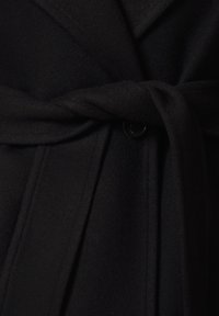 Czarny wełniany płaszcz z szerokim paskiem związanym w supeł wokół talii, z pojedynczym widocznym czarnym guzikiem pod paskiem.