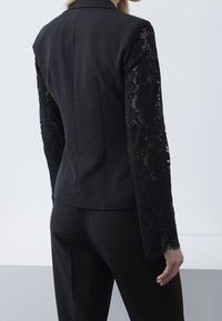 Mujer vistiendo un blazer negro ajustado con mangas largas de encaje floral y pantalones negros a juego, de pie contra un fondo claro y liso.