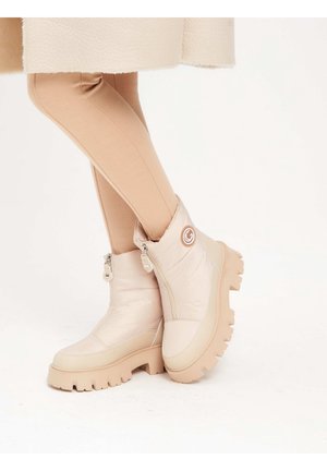 Jambes en leggings beige portant des bottines beige clair avec des semelles épaisses et des fermetures éclair latérales, debout sur un fond blanc uni.