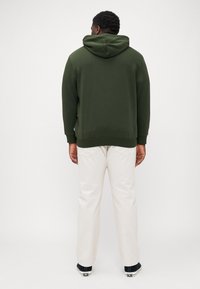 Green hooded sweatshirt with a smooth texture, worn over white pants. Sweatshirt features a kangaroo pocket and a ribbed hem.