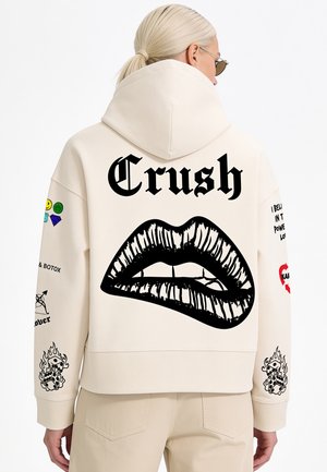 Person mit hellem Haar im Pferdeschwanz trägt einen cremefarbenen Hoodie mit schwarzer Grafik beißender Lippen und "Crush" Text auf dem Rücken sowie bunten Ärmelflicken.