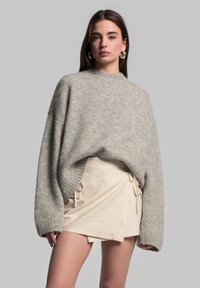 Übergroßer grauer Strickpullover mit weiten Ärmeln und hohem Kragen, kombiniert mit einem beigen Wickelrock mit seitlichen Bändern.