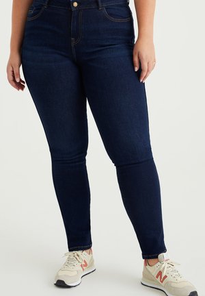 Jeans skinny blu scuro in denim, con cuciture chiare, vita alta e chiusura frontale con bottone. Abbinati a sneaker crema e rosse.