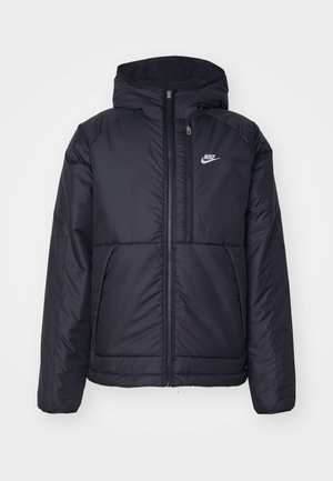 Black, padded Nike jacket with a front zipper and hood. Features a white logo on the left chest and dual front pockets. Smooth exterior texture.