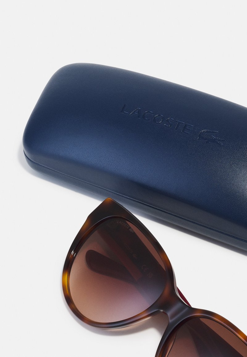 Lacoste zonnebrillen met bruin gradatie lenzen, oversized schildpadstijl monturen, liggen naast een marineblauwe hoes met reliëflogo.