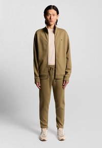 Tuta verde oliva con giacca con zip e pantaloni con cordino, realizzato in tessuto morbido. Abbinato a scarpe da ginnastica beige.