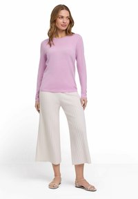 Hellrosa Langarmpullover kombiniert mit cremefarbenen gerippten, plissierten weit geschnittenen Hosen; das Model steht in Sandalen vor einem schlichten weißen Hintergrund.