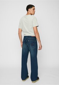 Lys grå t-skjorte kombinert med mørkeblå vide jeans. Jeansene har baklommer med unik sømdesign og en beige lærpatch.