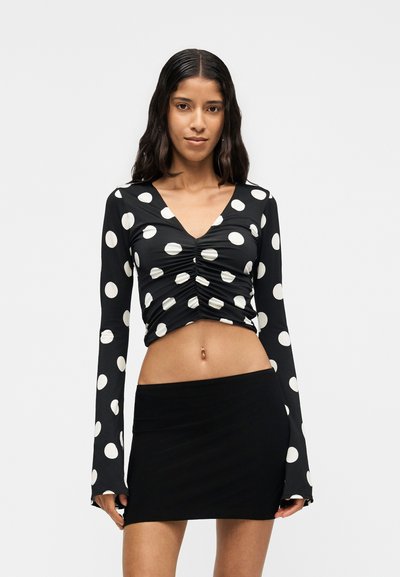 Γυναίκα που φοράει μαύρο crop top με μακριά μανίκια και μεγάλα λευκά πουά, καθώς και μαύρη μίνι φούστα, στέκεται μπροστά σε απλό φόντο.