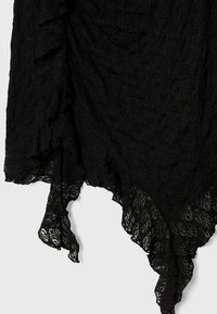 Tissu en dentelle noire avec un bord en forme de festons et un motif floral, suspendu devant un fond blanc.