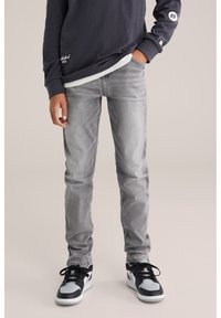 Sive jeans iz kašmira s slim fit, z detaljnim šivanjem in blede zaključne obdelave, v kombinaciji z črnimi in belimi visokimi športnimi copati.