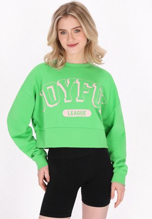 Ung kvinde iført en lys grøn cropped sweatshirt med "JOYFUL LEAGUE" trykt på fronten og sorte tætsiddende shorts, smilende med hånd på hoften.