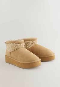 Tan ruskind støvler til anklerne med en fluffy cremeforing, rund tå og en tyk tan platformsål. Dekorativ pull tab bagpå.