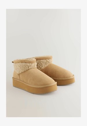 Vaaleanruskeat mokkanahkaiset nilkkasaappaat, joissa on pörröinen kerma-valkoinen vuori, pyöristetty kärki ja paksu vaaleanruskea alustalla. Koristeellinen vetoketju takana.