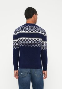 Maglione blu navy con motivo geometrico in crema e beige. Tessuto a maglia, scollatura rotonda, polsini e orlo a costine. Indossato con jeans blu.