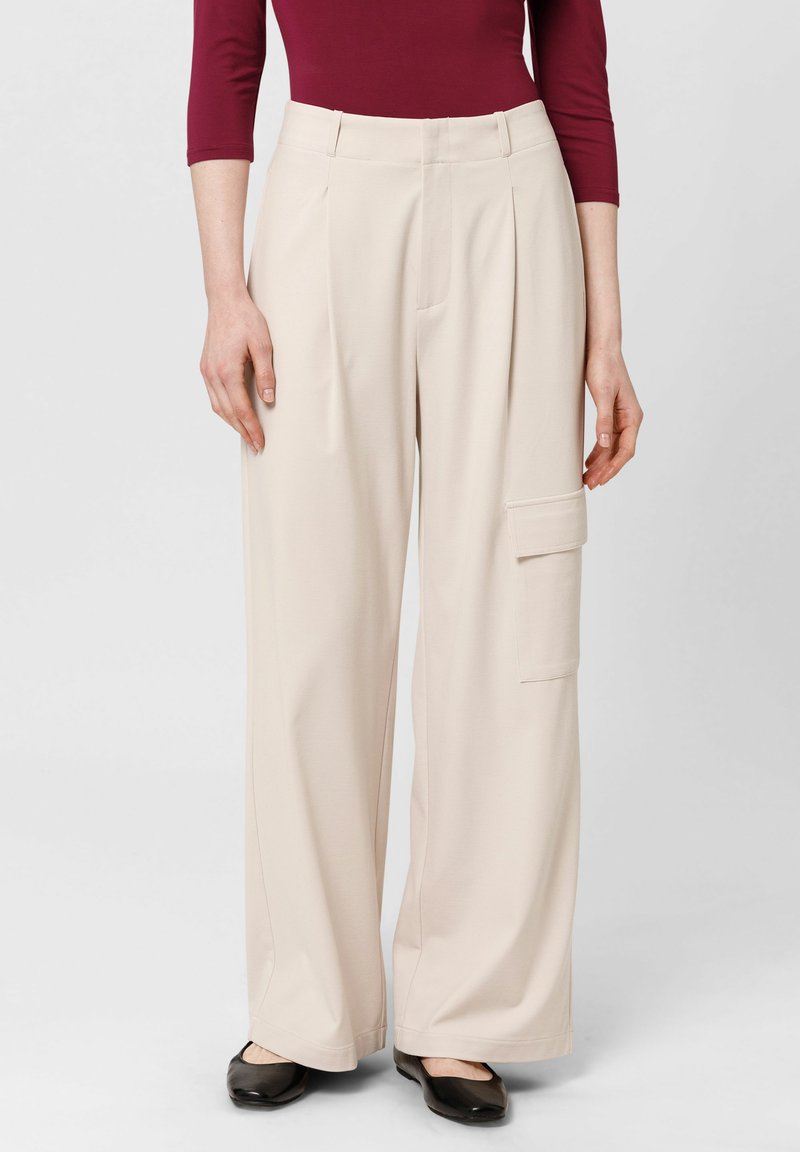 Beige wijde cargobroek met plooien, zijkleeddetails en een gladde, zachte stof. G gedragen met zwarte platte schoenen en een bordeauxrood top.