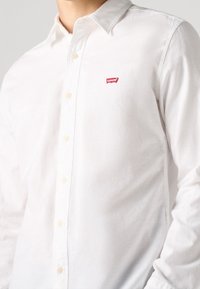 Weißes Baumwoll-Hemd mit Struktur, das vorne ein kleines rotes Levi's-Logo und beige Knöpfe aufweist.