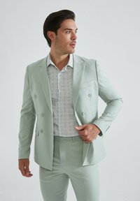 Costume double-breasted vert clair avec un pantalon assorti, associé à une chemise grise texturée avec de petits motifs et un col boutonné.