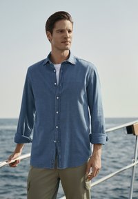 Camicia in chambray blu con maniche lunghe, colletto button-down e vestibilità rilassata. Indossata su una t-shirt bianca, abbinata a pantaloni cargo color oliva.