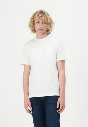 White cotton t-shirt featuring a textured "GUESS" logo in the center. Short sleeves and a round neckline; paired with dark blue jeans.