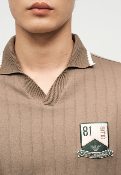 Κοντινό πλάνο ενός ατόμου που φορά μια μπεζ πόλο μπλούζα με ριμπ λαιμόκοψη σε σχήμα V και ένα κεντητό έμβλημα Emporio Armani 81 Club.