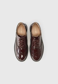 Burgundy lackläder skor med broguedetaljer, runda tår och bruna snörningar. Inkluderar en texturerad svart sula.