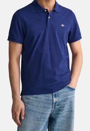 Homme portant un polo bleu à manches courtes avec un petit logo sur la poitrine et un jean bleu clair, main dans la poche.