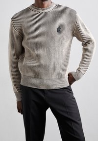 Gestrickter Pullover mit vertikalen schwarzen und beigen Streifen, rundem Hals und gerippten Bündchen. Hat ein kleines schwarzes Logo auf der Brust.