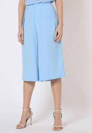 Mujer vestida con pantalones anchos de pierna corta de color azul claro y sandalias de tacón alto rosa pálido, de pie frente a un fondo liso.