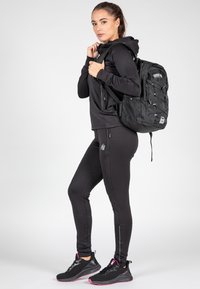 Svart sportig outfit som inkluderar en åtsittande hoodie och leggings, kombinerad med en svart ryggsäck med meshdetaljer och justerbara remmar.