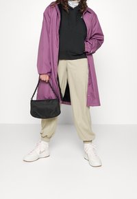 Abrigo de lluvia morado de gran tamaño sobre una sudadera negra con capucha, pantalones de chándal beige y zapatillas blancas. Sosteniendo un bolso negro con una correa de cadena.