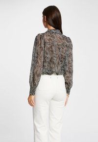 Zwart-wit patroonde blouse van transparant materiaal met pofmouwen en knoopmanchetten, gecombineerd met hooggeritste witte broeken.