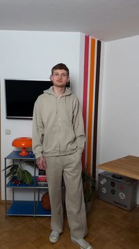 Νέος άνδρας με μπεζ φούτερ με κουκούλα και παντελόνι στέκεται σε δωμάτιο με ριγωτό τοίχο, μπάλα του μπάσκετ, πορτοκαλί λάμπα και μεταλλικό ράφι με φυτά και βιβλία.