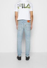 Tylny widok osoby w białej koszulce z kolorowym logo FILA oraz jasnoniebieskich dżinsach z czerwoną łatą Levi's i dwoma tylnymi kieszeniami.