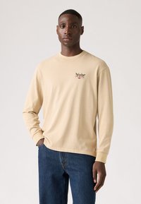 Beige langwerpige t-shirt met lange mouwen en een ronde hals, voorzien van een klein grafisch ontwerp op de borst. Zachte katoenen stof, relaxte fit, gecombineerd met blauwe spijkerbroek.