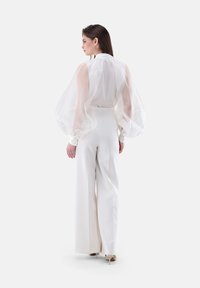 Blusa bianca trasparente con maniche a sbuffo voluminose abbinata a pantaloni bianchi a vita alta e tacchi nudi, vista da dietro.