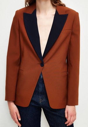 Vrouw draagt een roestbruin colbert met brede zwarte revers en knoop, gecombineerd met donkerblauwe jeans, tegen een effen lichte achtergrond.