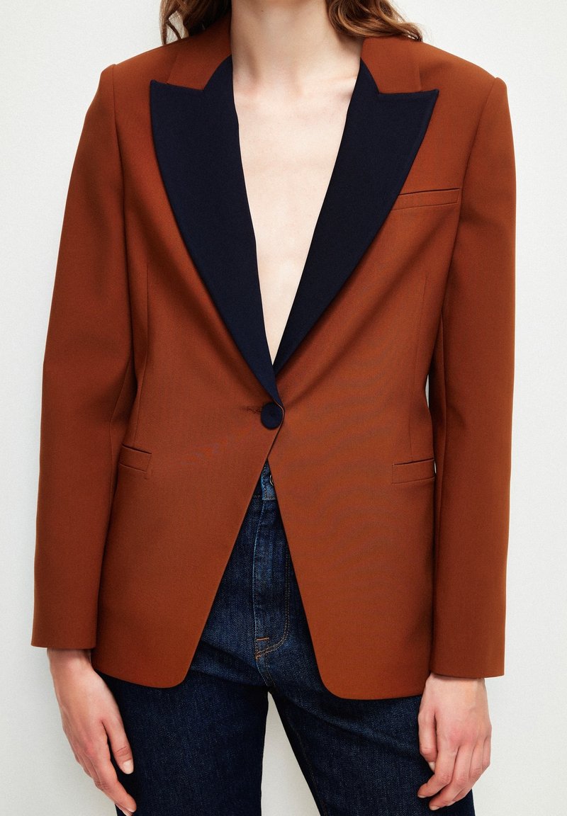 Femme portant un blazer marron rouille avec de larges revers noirs et un bouton, associé à un jean bleu foncé, sur un fond clair uni.