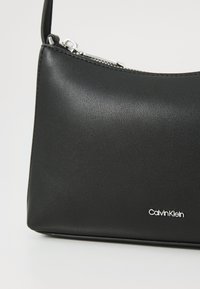 Gentă de umăr din piele neagră cu o textură netedă, închidere cu fermoar și accesorii din argint. Prezintă logo-ul "Calvin Klein" în argint.