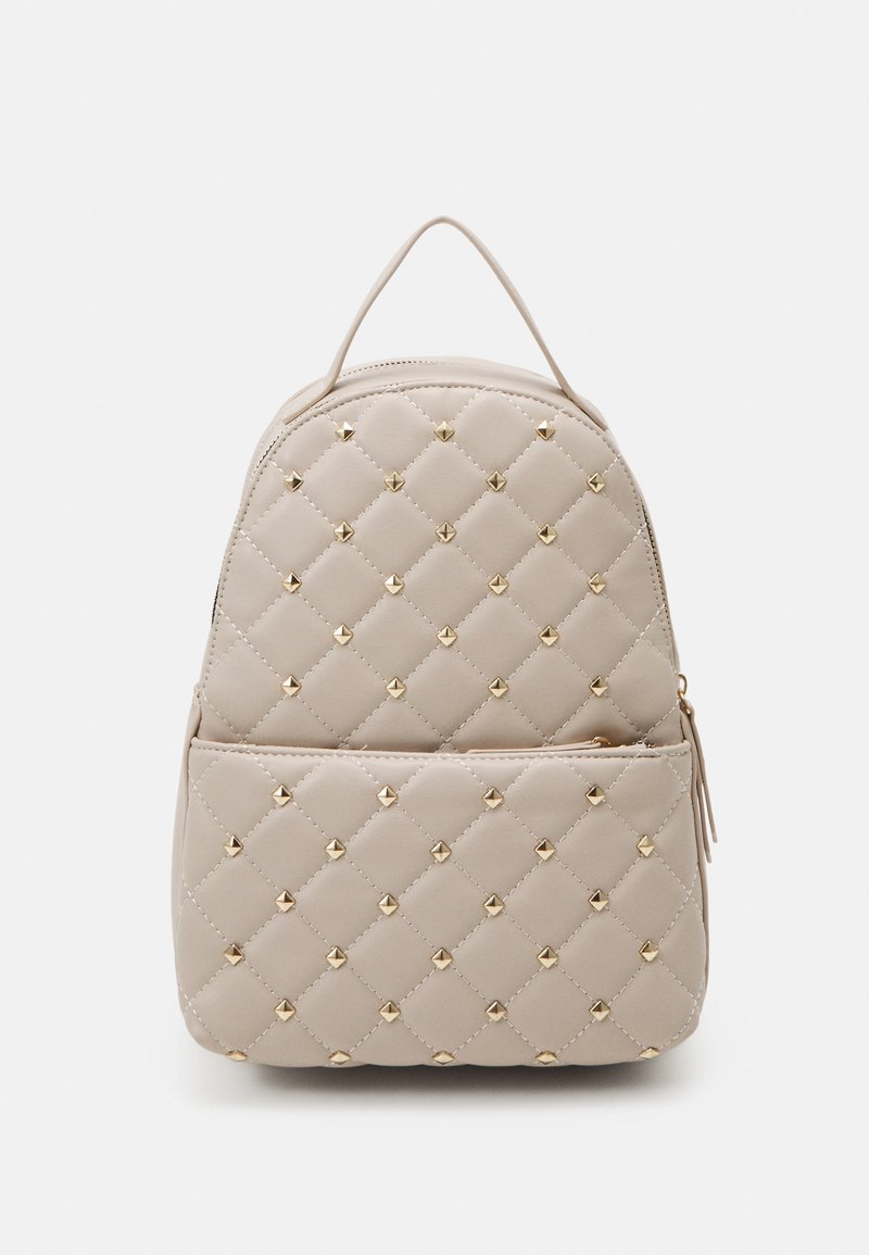 Petit sac à dos matelassé beige avec des clous dorés, une poche avant zippée et une poignée supérieure sur un fond clair uni.