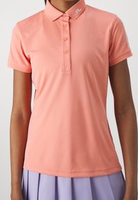 Kvinna i en korallrosa kortärmad polo med knappar och en lavendelfärgad plisserad kjol, sedd från halsen till höfterna.