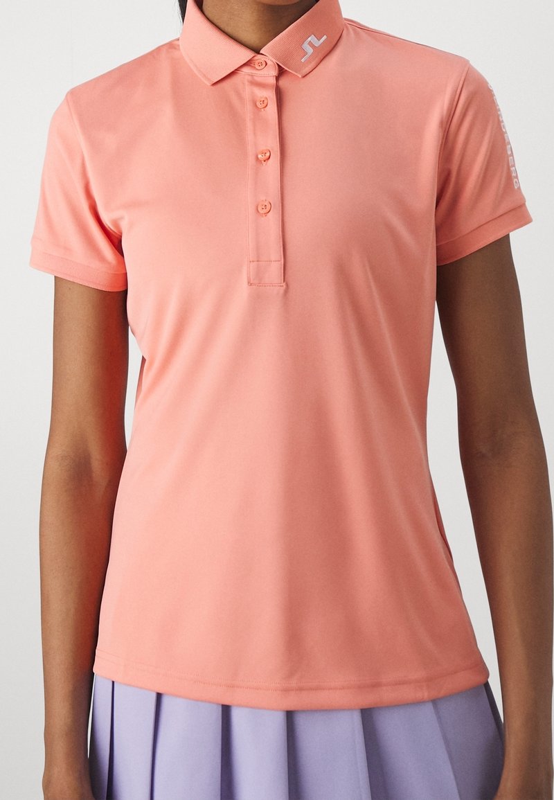 Kvinna i en korallrosa kortärmad polo med knappar och en lavendelfärgad plisserad kjol, sedd från halsen till höfterna.