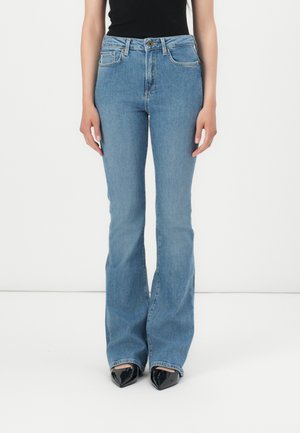 Mulher vestindo jeans de cintura alta em corte bootcut azul-claro e sapatos pretos de bico fino contra um fundo branco.