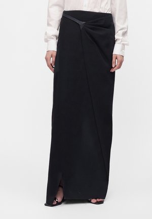 Femme portant une longue jupe portefeuille noire avec une fente frontale, associée à des escarpins ouverts noirs et une chemise blanche boutonnée.